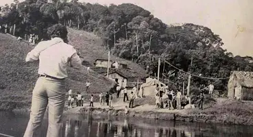 Foto histórica de um jovem sobre uma embarcação na margem do Rio Juquiá, olhando para uma multidão reunida em frente a ranchos de palha e a encosta do Morrinho.