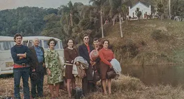 Foto colorida de Julho de 1971 de um grupo de pessoas com malas e uma Kombi, reunidos na beira do rio, com o Templo de Morrinho visível ao longe na colina.