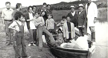 Foto histórica em preto e branco de um grande grupo de famílias (incluindo crianças e adultos), em volta de um barco de madeira, na margem do Rio Juquiá, com morros e vegetação ao fundo.