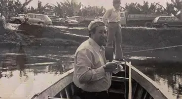 Foto antiga do Rev. Evandro Luis em um barco de madeira no rio, com carros e caminhões estacionados na margem, simbolizando a transição do acesso para o Morrinho.