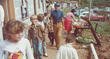 Foto colorida de 1982 mostrando crianças e adultos em um caminho de terra no Morrinho. Uma mulher prepara alimentos em uma mesa rústica ao lado de ranchos, e carros estão estacionados ao fundo.